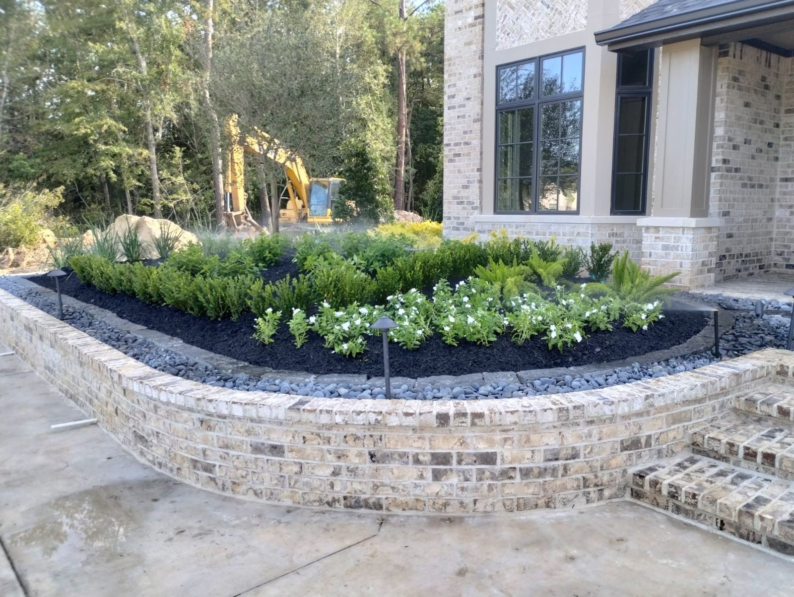 Brick planter with greenery in front of a house, with a small excavator in the background.