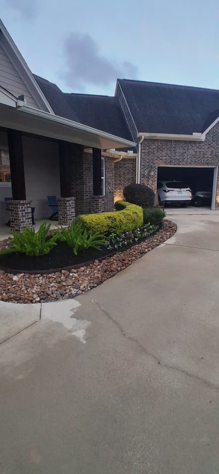 A stone house with a covered porch and a landscaped garden bed containing shrubs and ferns, alongside a concrete driveway.