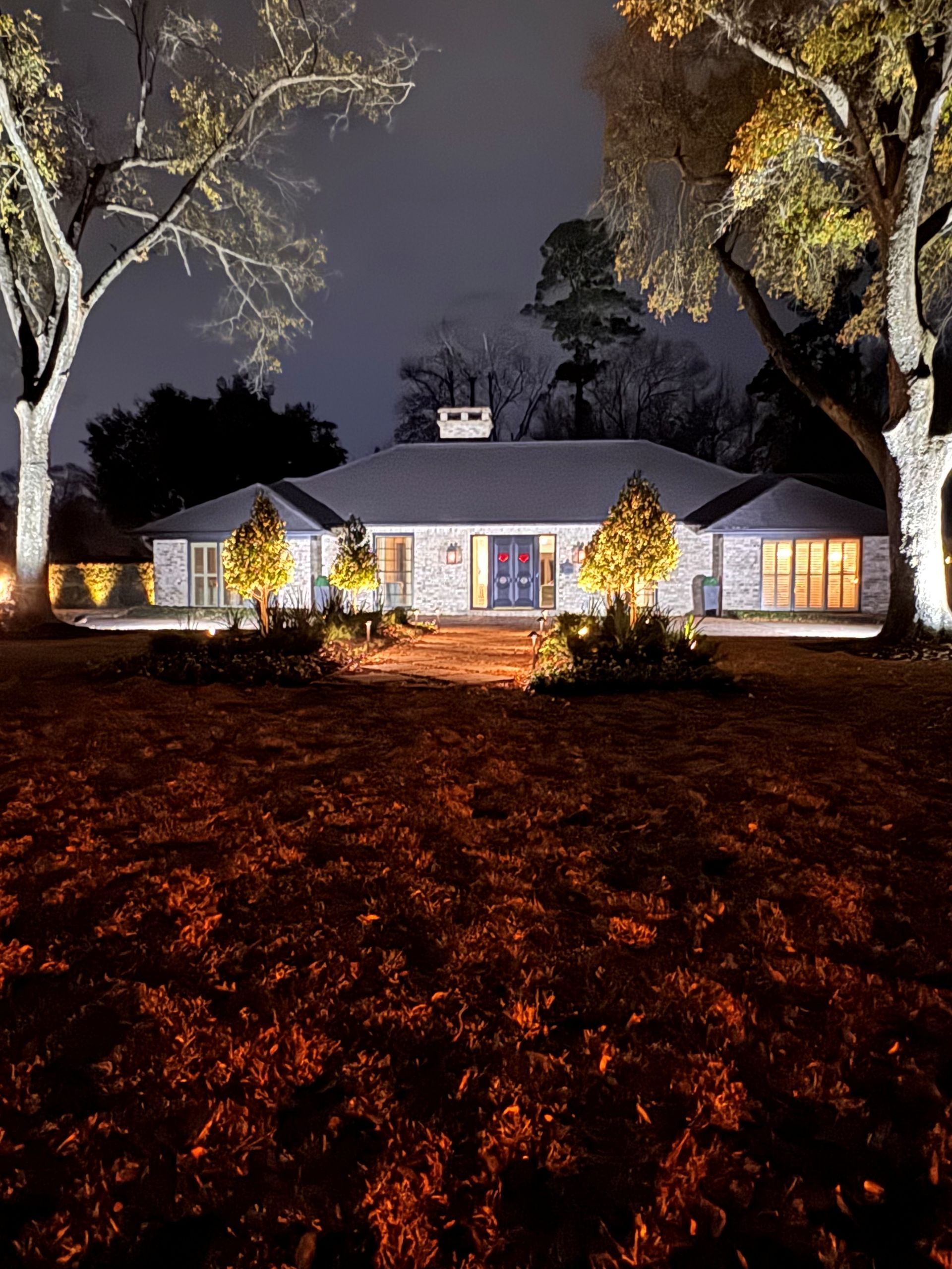 Night view of a house with landscape lighting illuminating the facade, trees, and lawn.