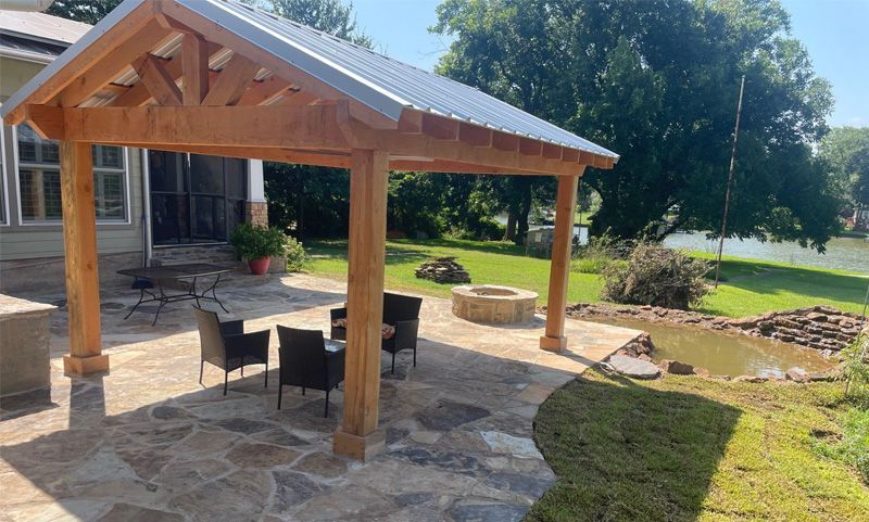 A wooden pavilion with a metal roof on a patio with fire pit in the backyard of a house