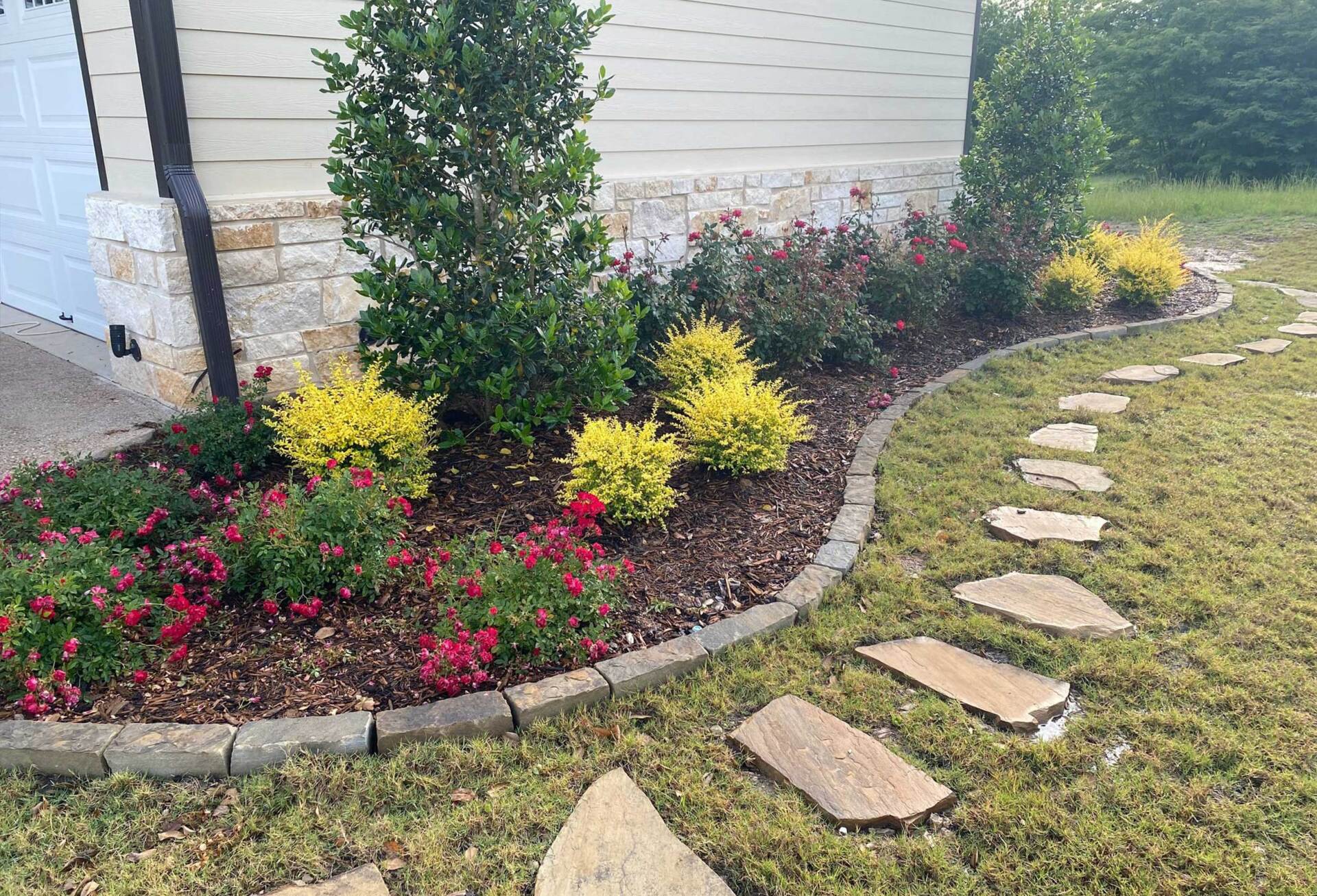 A stone walkway leading to a house surrounded by flowers and bushes.