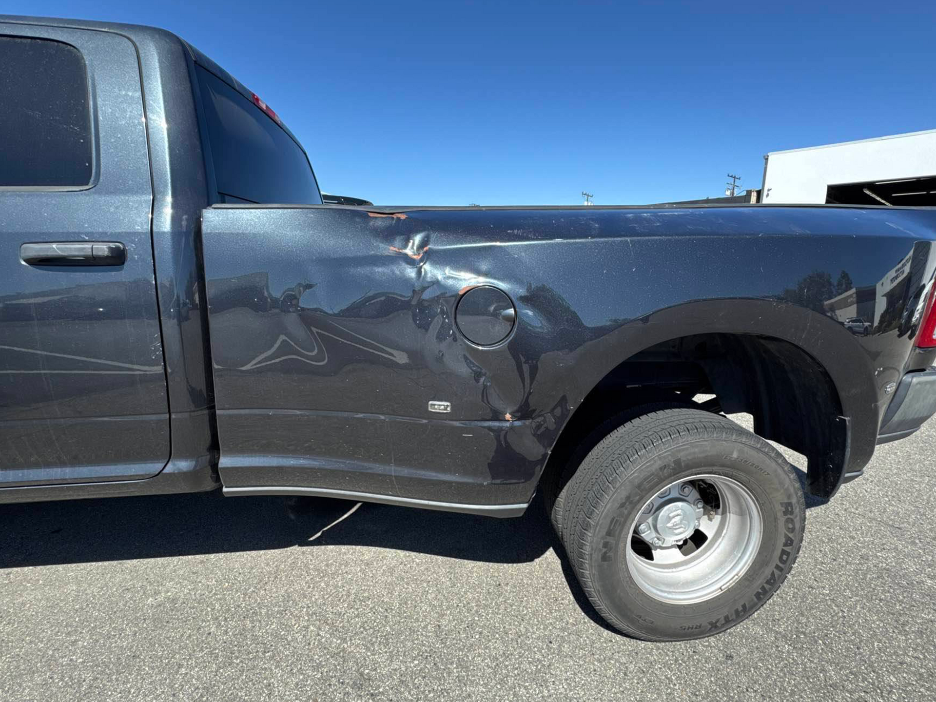 Damaged black pickup truck with a dented side panel and rear wheel visible, parked outdoors.