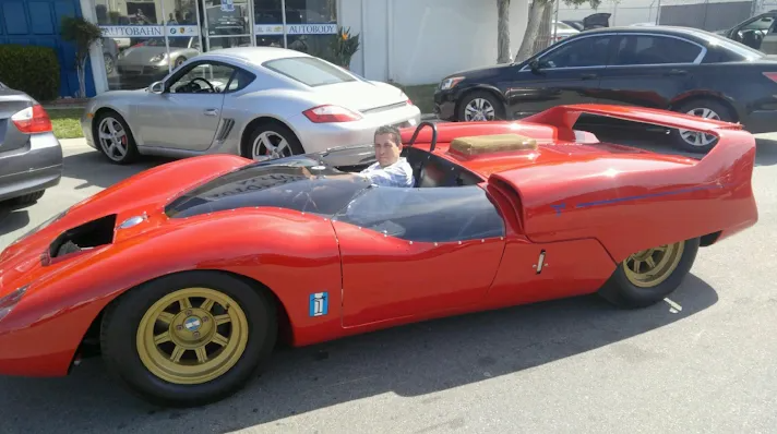 Red vintage race car parked on a street with a person inside; gold wheels, black windshield, sunny day.