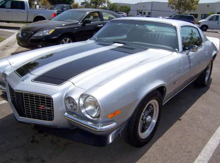 Silver 1970s Chevrolet Camaro with black racing stripes parked outside in a sunny area.