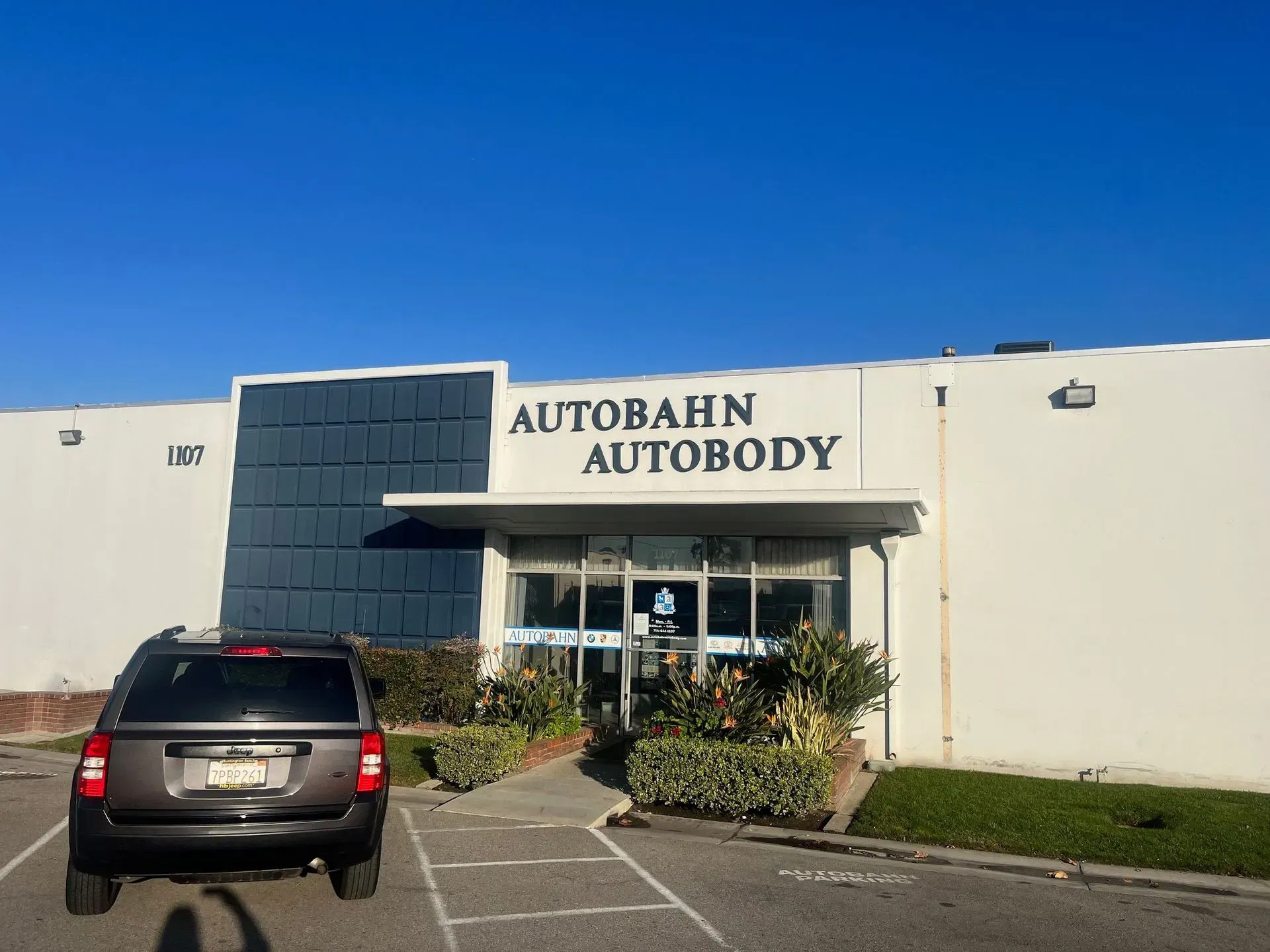 Autobahn Autobody shop exterior. Gray SUV parked outside white building with blue accents. Clear blue sky.