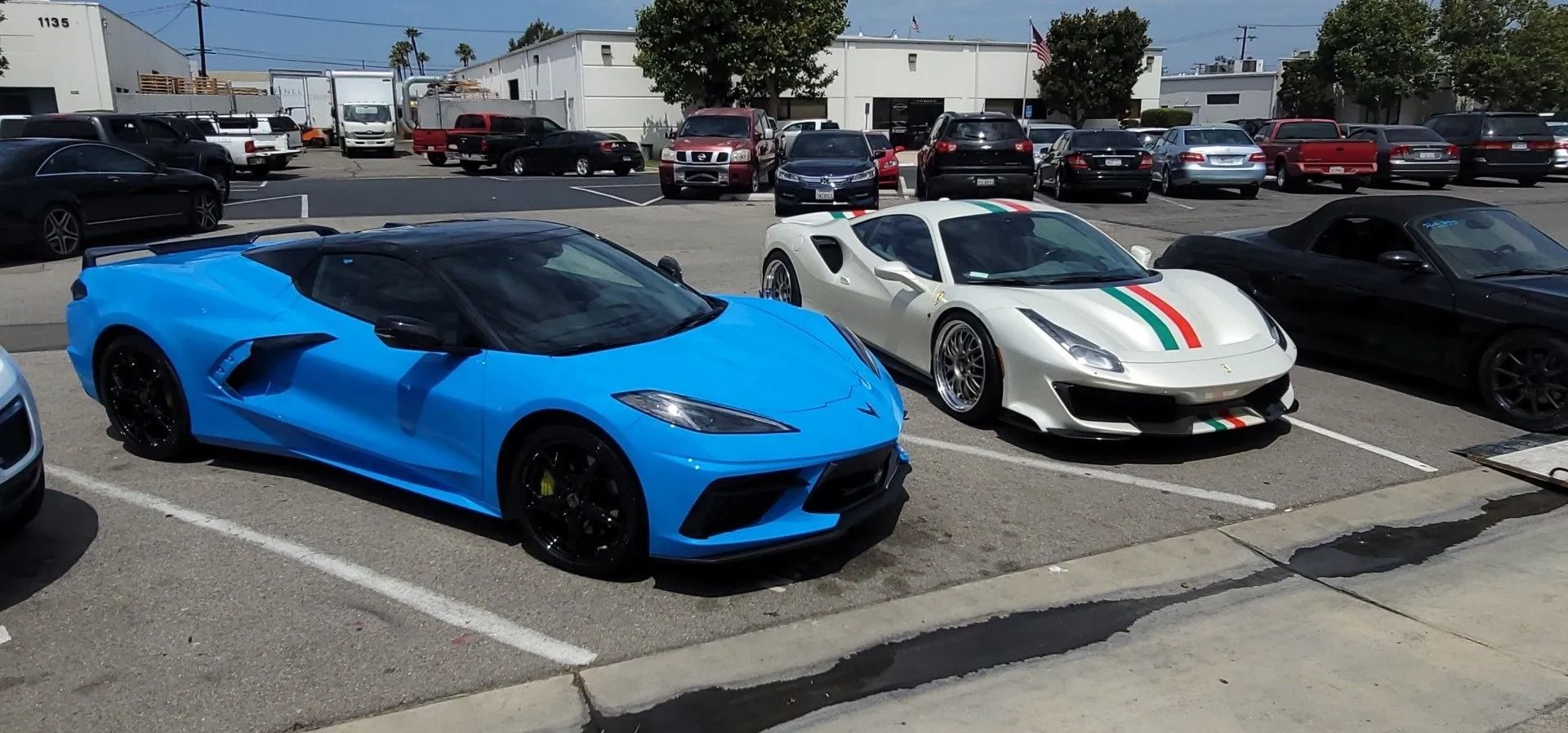 Blue sports car parked next to a white sports car with green/red stripe and other vehicles in a parking lot.