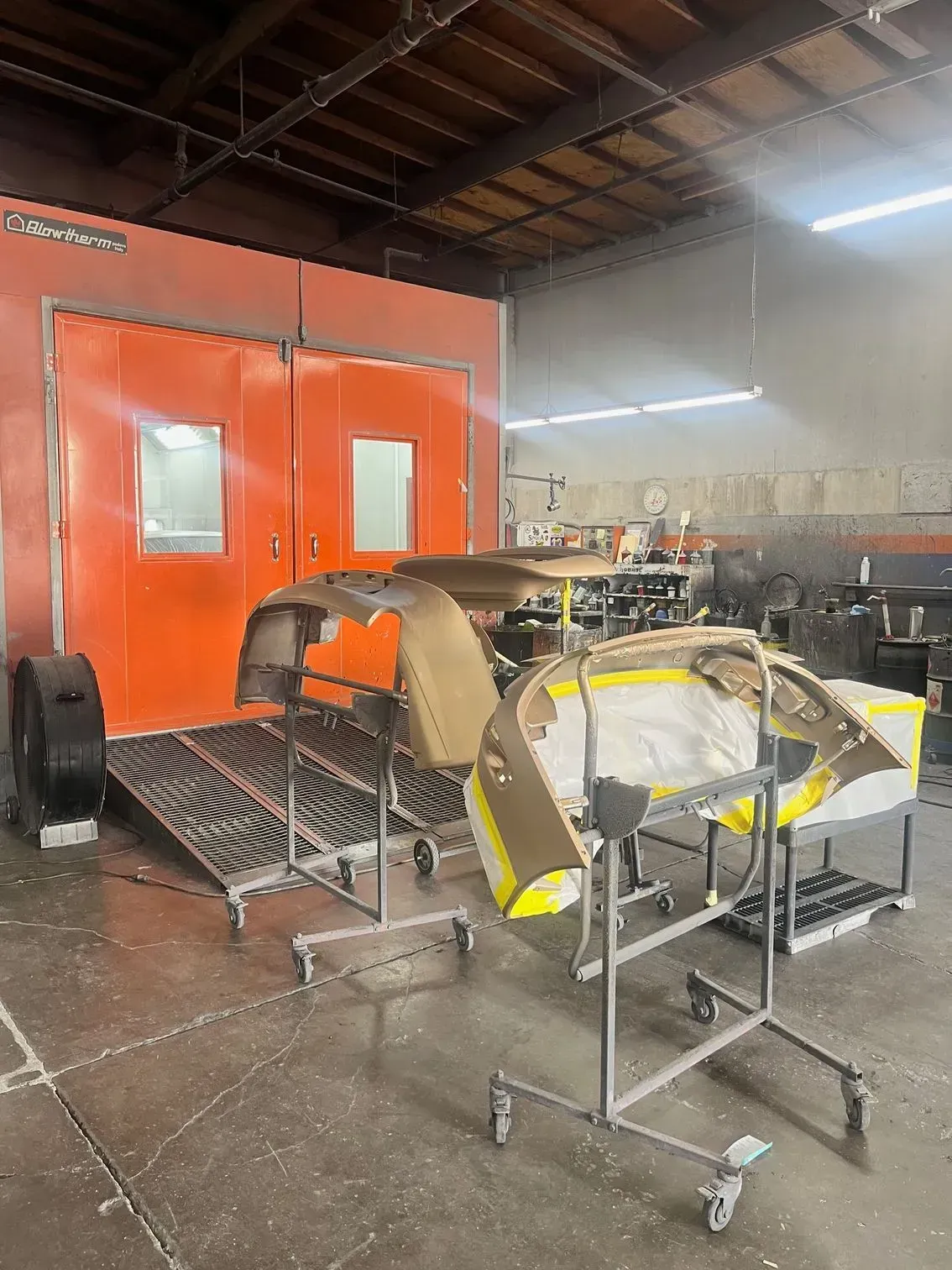 Car parts on stands in a body shop, orange paint booth in the background.