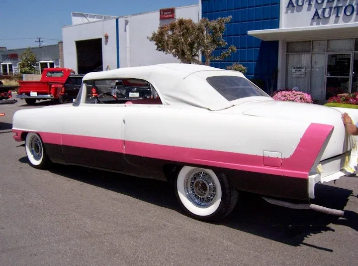 White and pink vintage car parked on a street in front of a blue building, with a red truck in the background.
