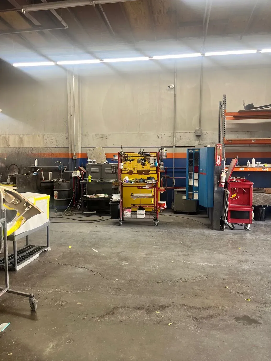 Interior of a cluttered auto repair shop, with tools, equipment, and shelves against concrete walls and floor.