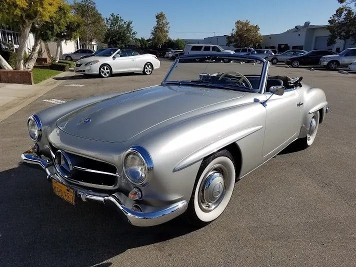 Silver vintage Mercedes-Benz convertible parked on asphalt. White-walled tires, chrome accents, and a clear blue sky.