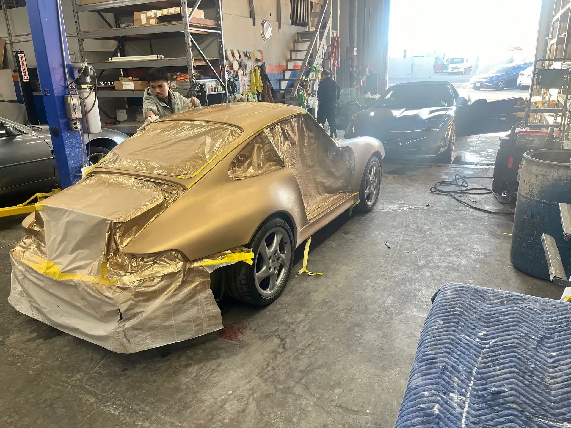 Gold-colored Porsche in a garage, taped and covered for painting. Other cars visible nearby.