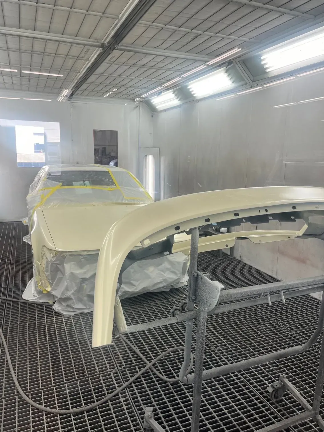 Car in a paint booth, partially masked. A painted fender sits in the foreground. Creamy yellow.