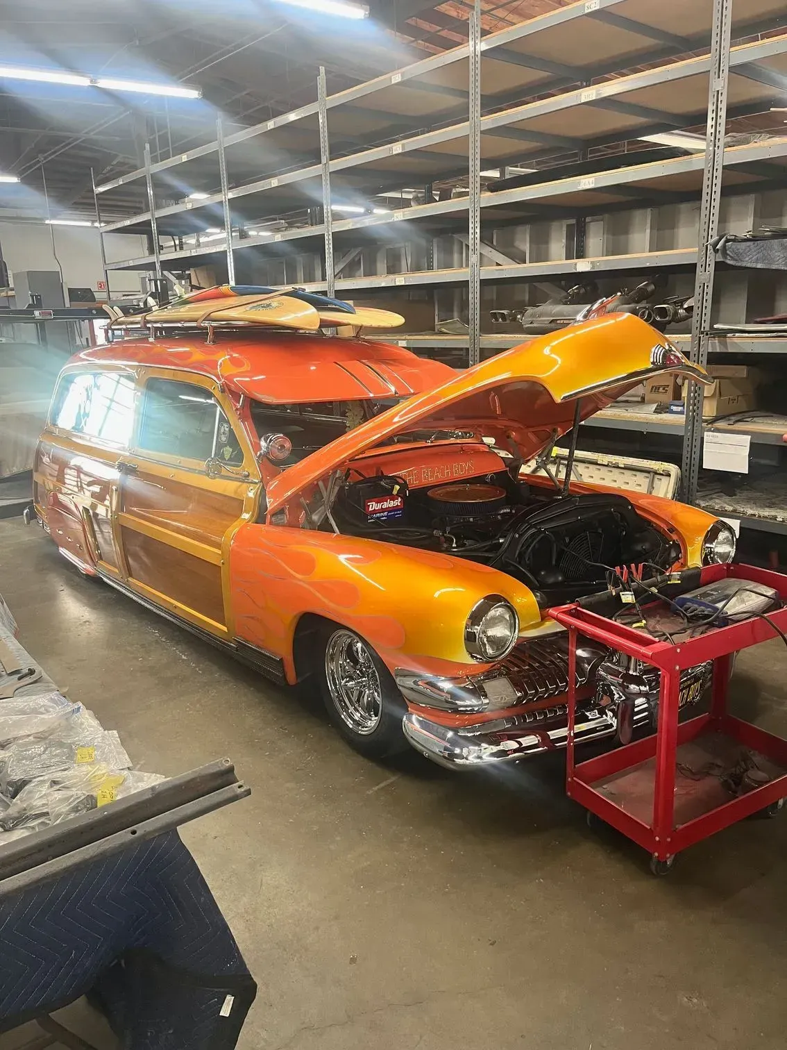 Orange custom woodie wagon with hood open, surfboards on top, parked in a workshop.