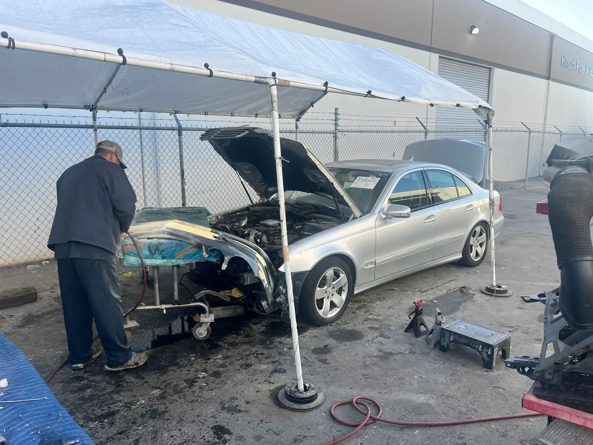 Car damaged, under repair. Person working on it under a canopy, outdoors. Silver sedan.