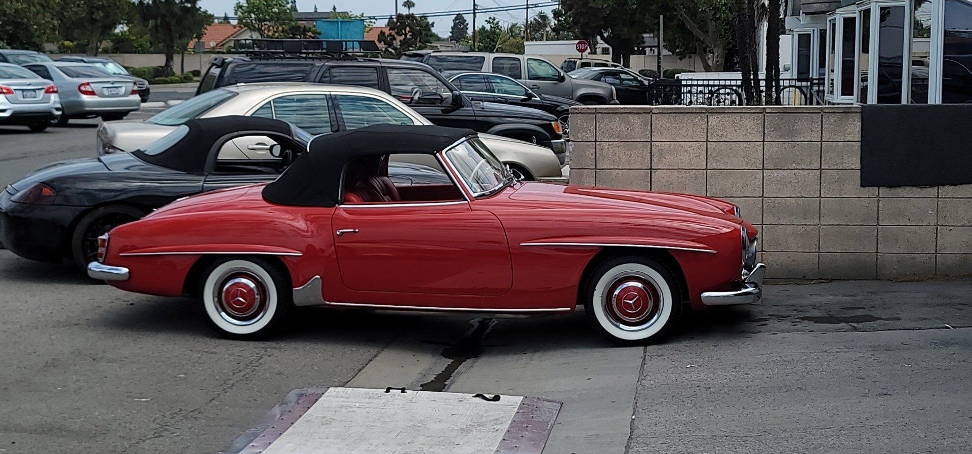 Red vintage convertible with white-walled tires parked on a paved lot. Black car next to it, other cars in background.