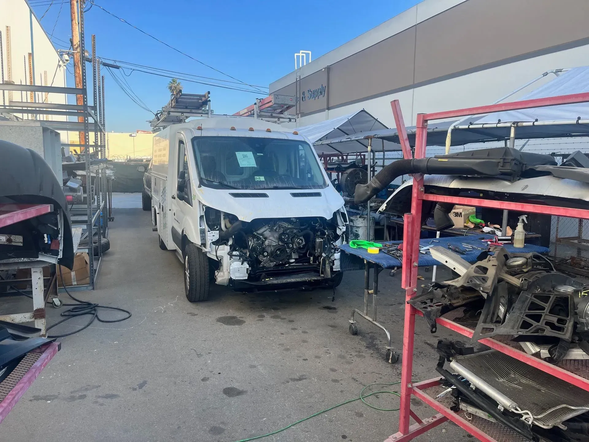 White van with front damage in a salvage yard, surrounded by parts and equipment.