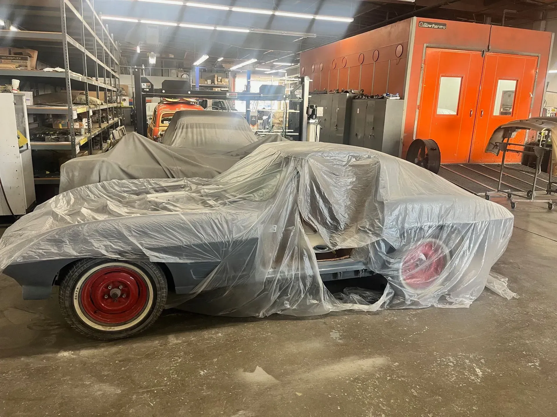 A classic car covered in plastic in a garage, red wheels visible, orange paint booth in the background.