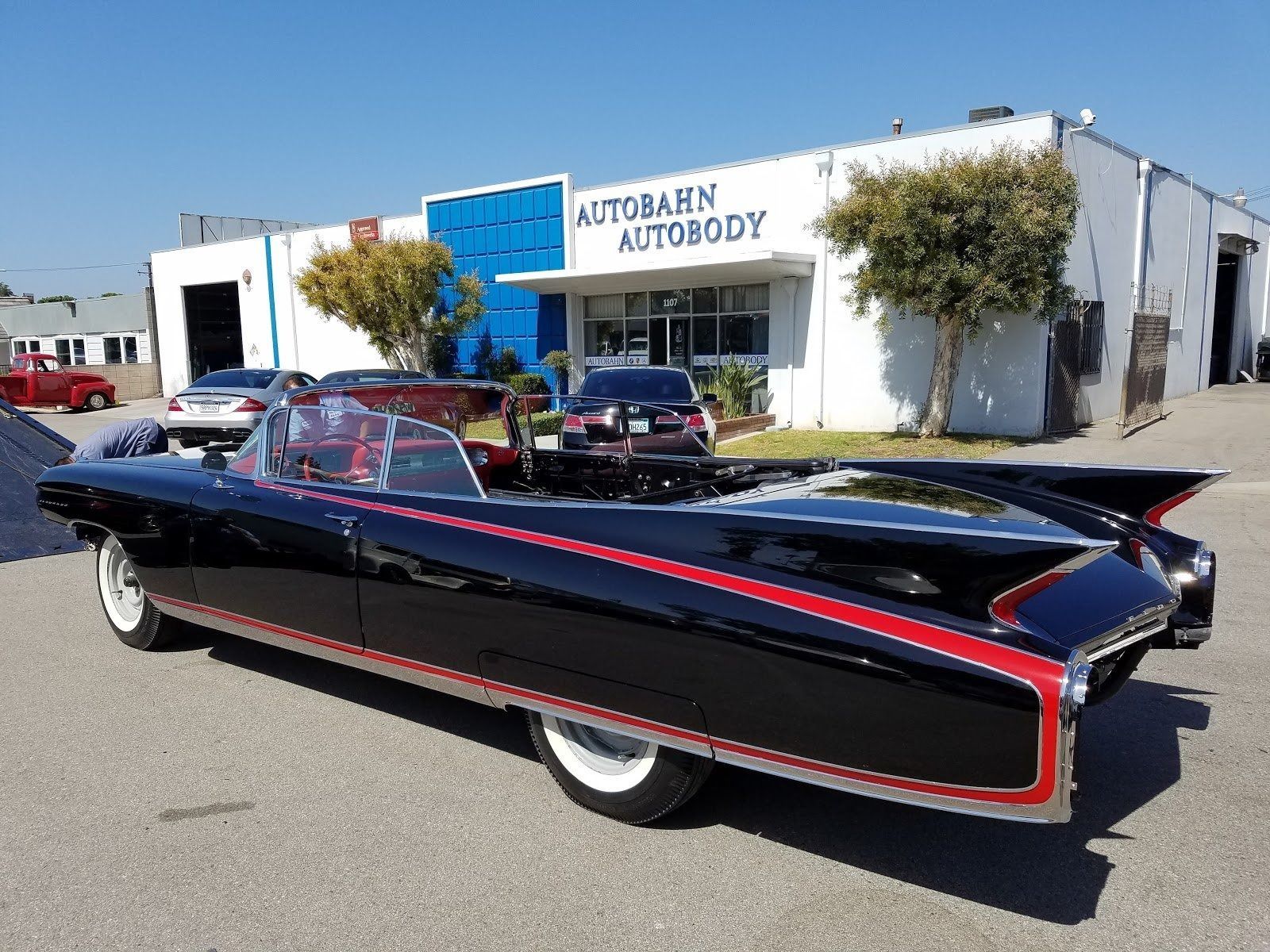 Black custom car with red trim parked in front of autobody shop on sunny day.