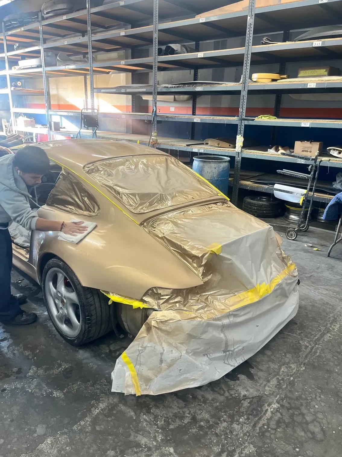 A person in a workshop wiping down a gold-painted Porsche. The car is partially covered in masking.