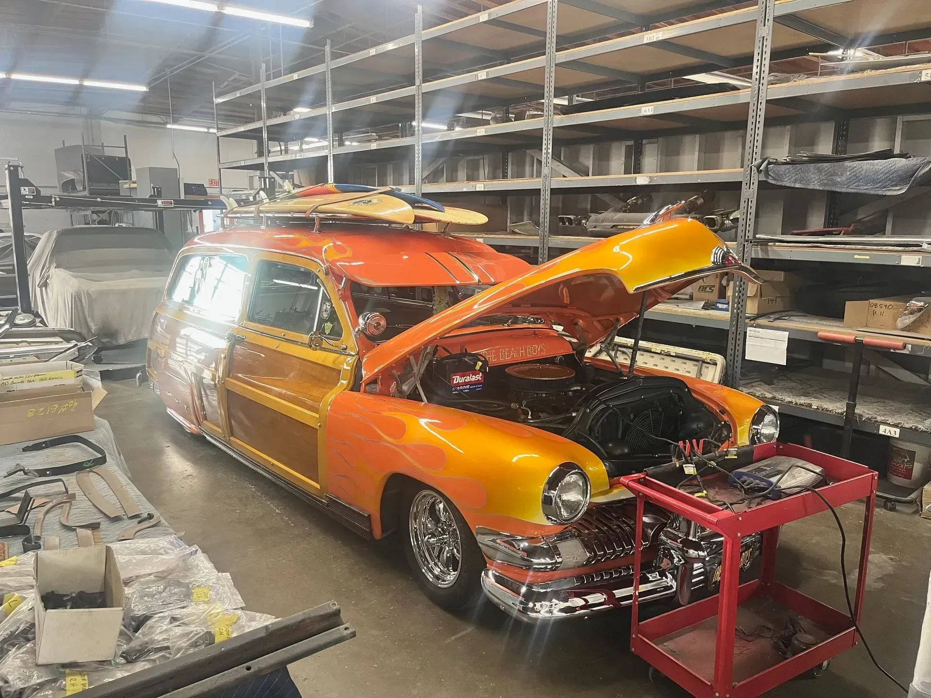 Orange and wood-paneled classic car with surfboard on top, hood open, in a workshop.