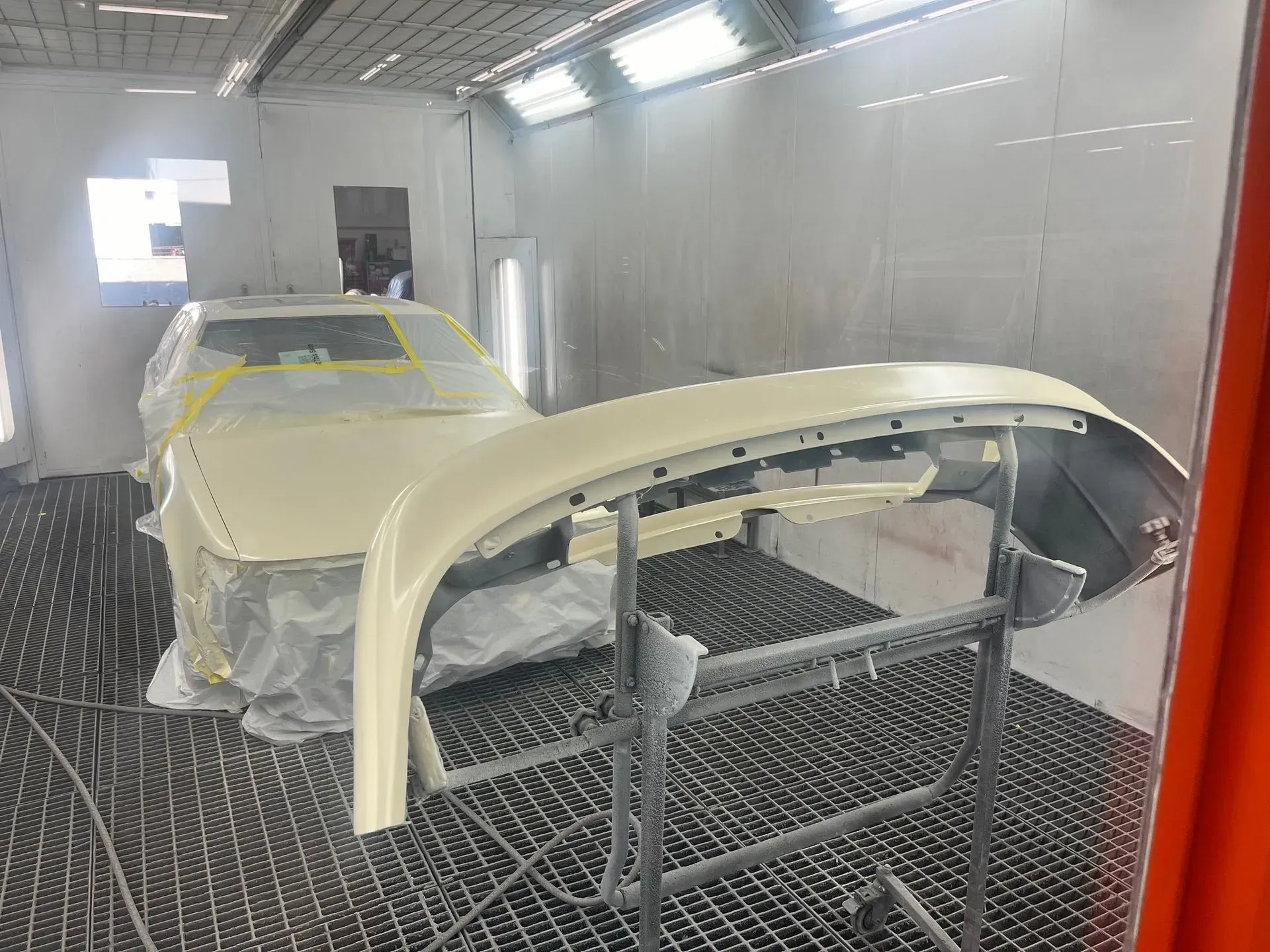 Car bumper and vehicle body in a paint booth, covered in plastic, ready for painting.