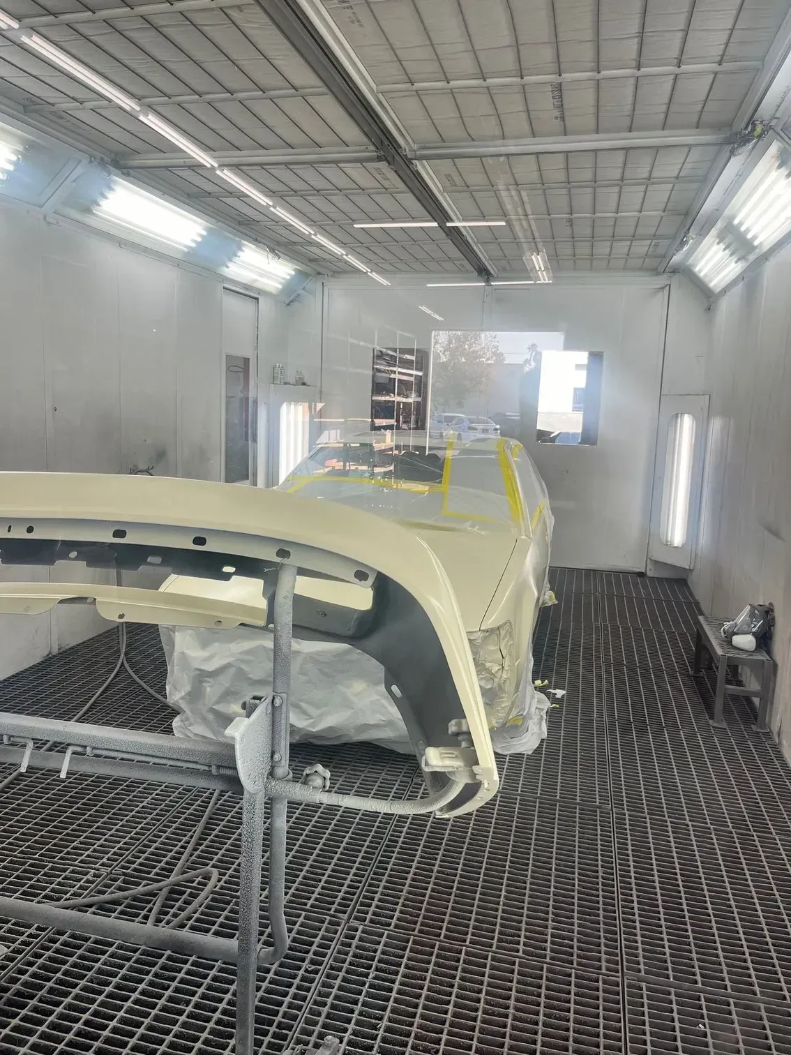 Inside a paint booth, a vehicle is partially covered, with a bumper on a stand.
