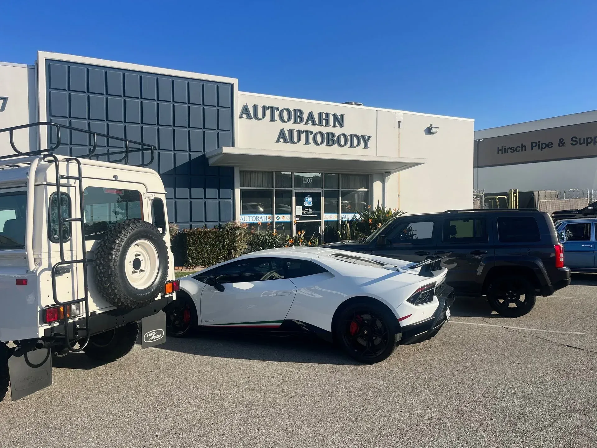 White Lamborghini parked outside Autobahn Autobody shop. Other vehicles also present. Bright day.