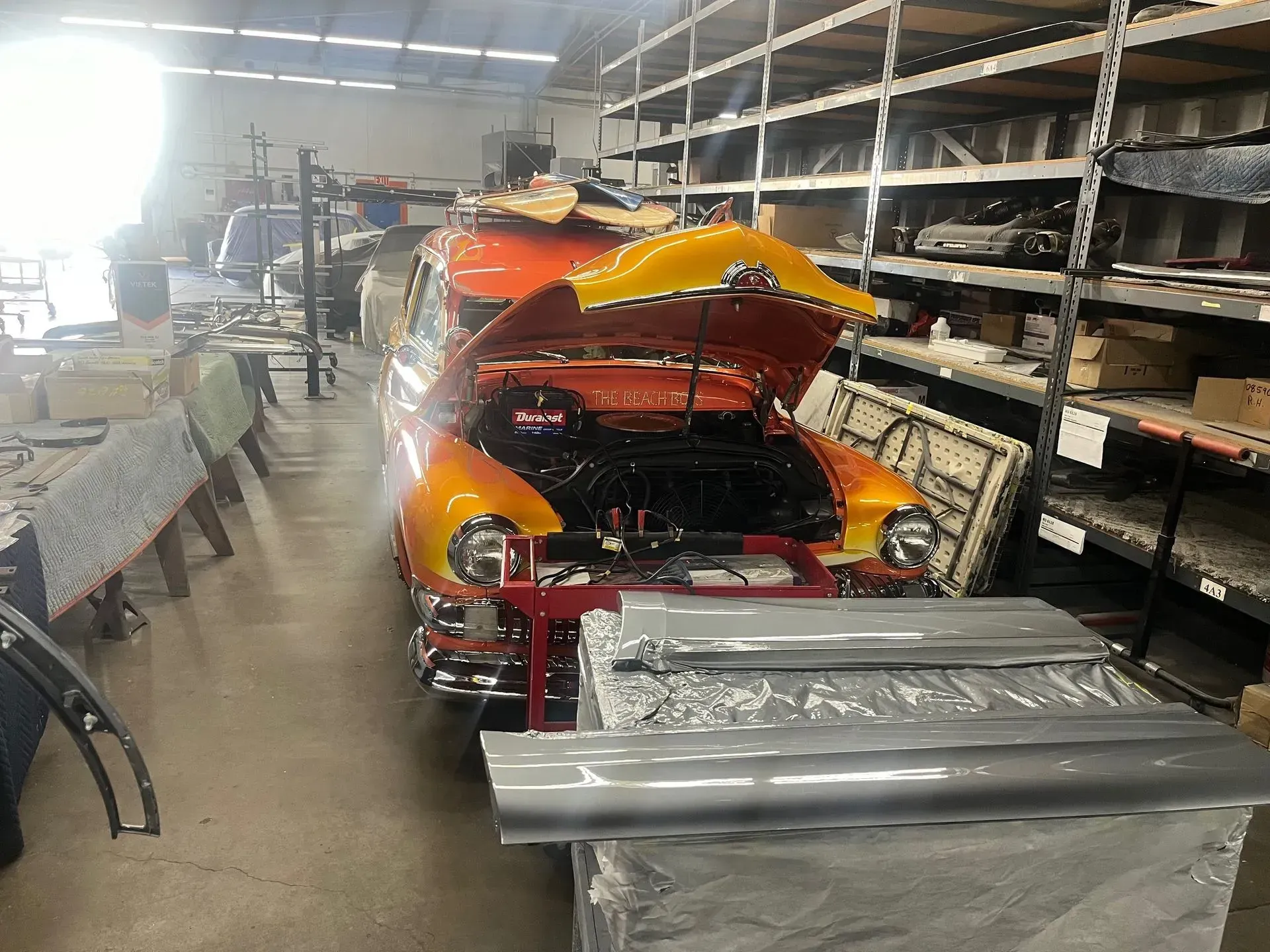 Classic orange car with open hood in a warehouse, parts on shelves and table.
