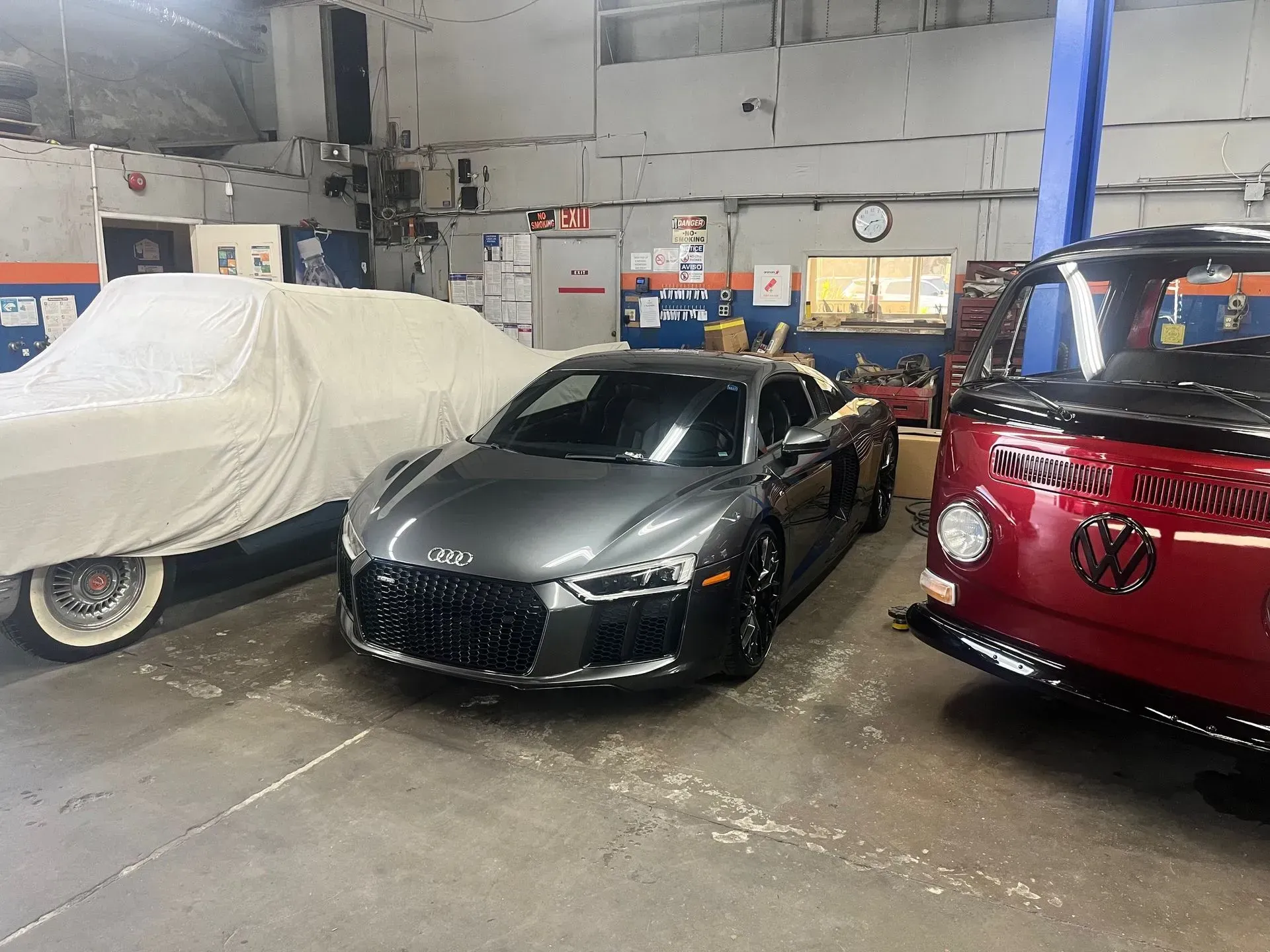 A gray Audi R8 sports car parked in an auto repair shop next to a covered car and a red VW bus.