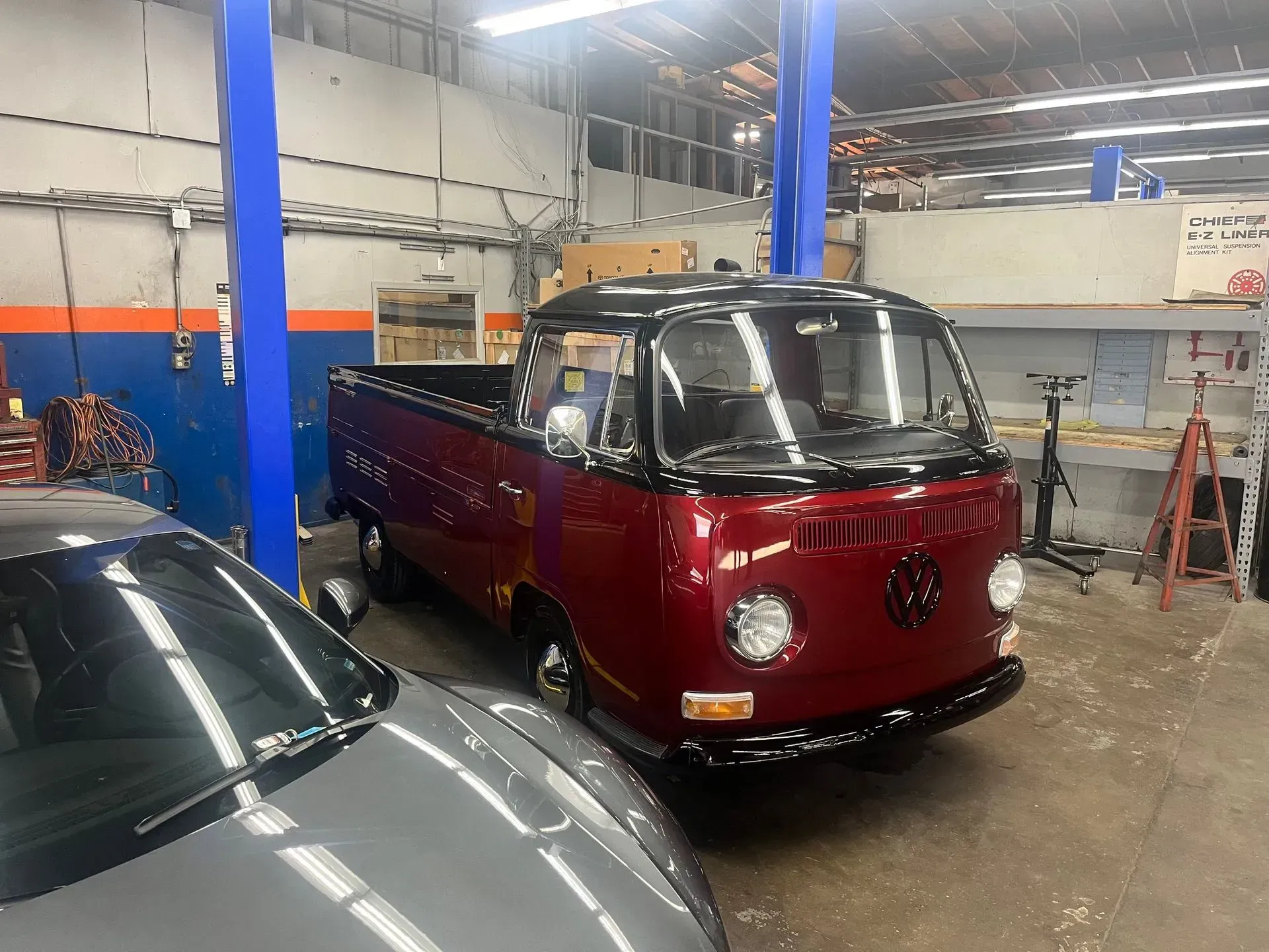 Burgundy and black Volkswagen pickup truck inside a mechanic's garage, parked near a silver car.