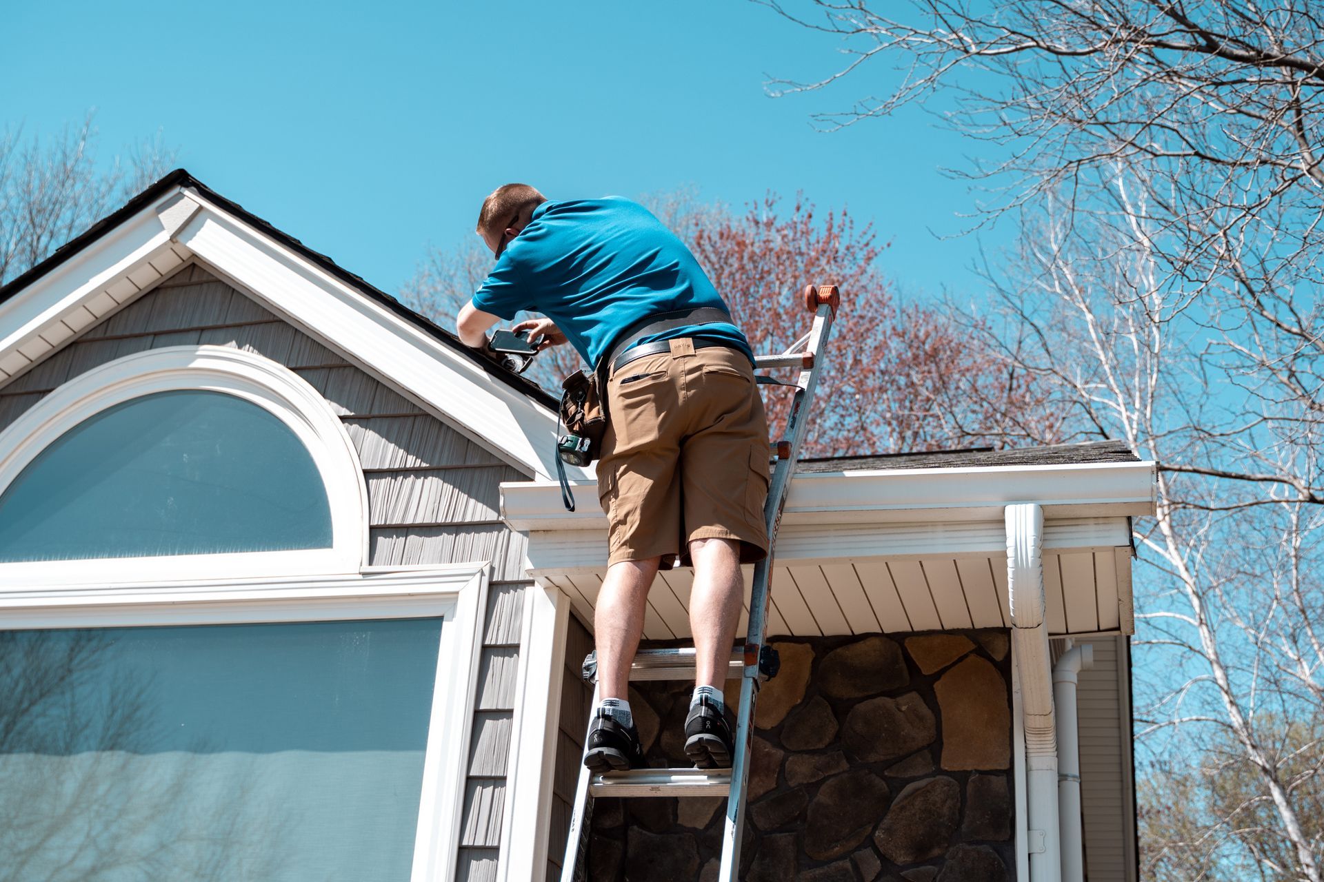 Person on ladder, working on house gutters, blue shirt, tan shorts, sunny day.