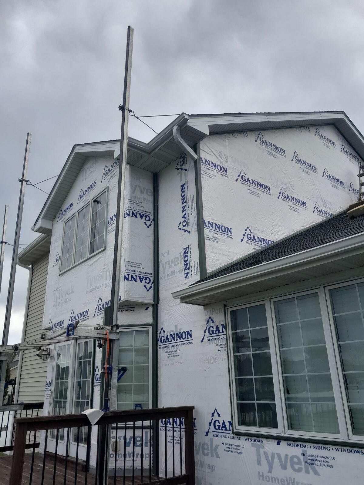 House under construction, covered in Tyvek wrap, scaffolding visible, gray sky background.