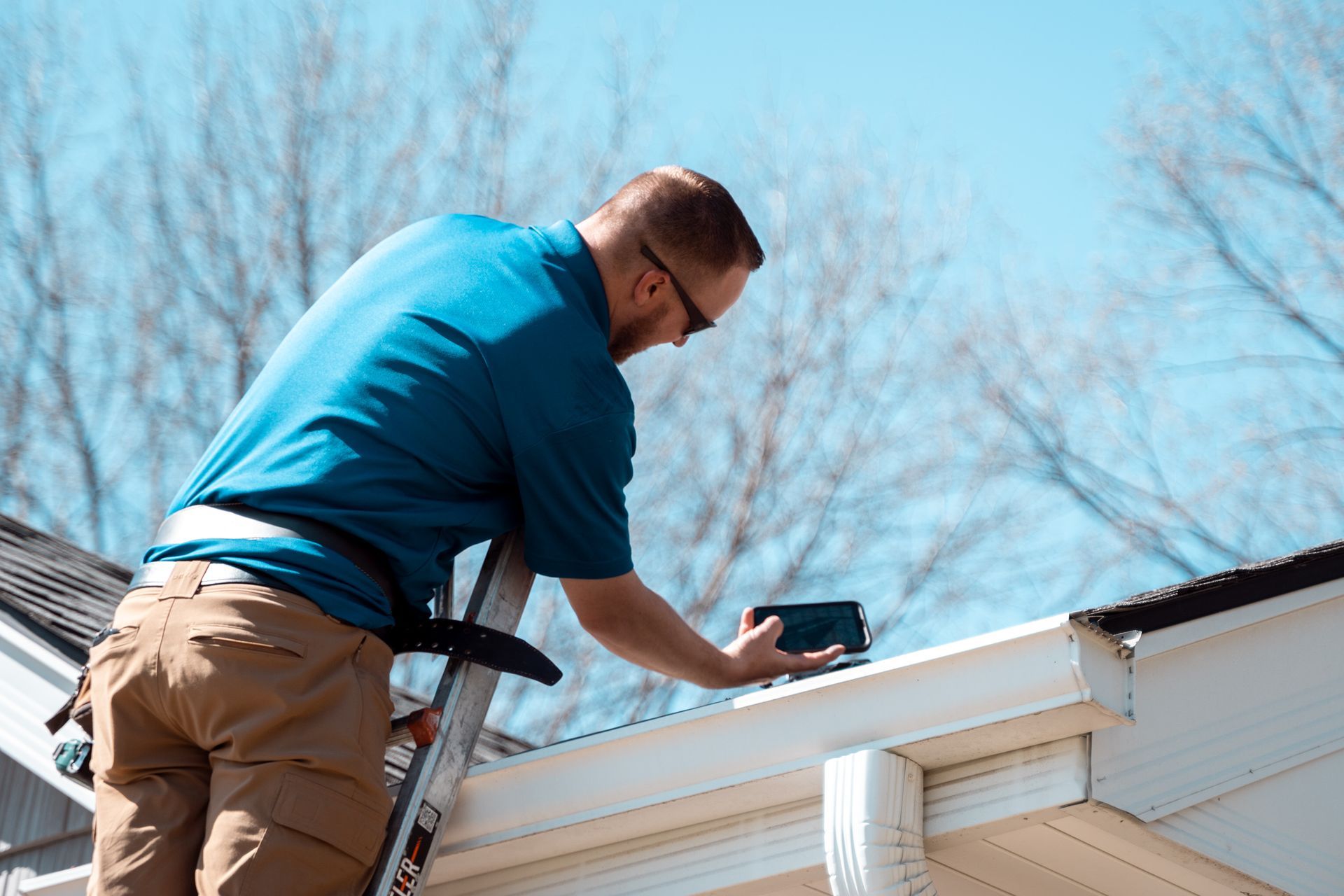 Man on a ladder inspecting a white gutter, holding a tablet. Sunny day, blue shirt, tan pants.