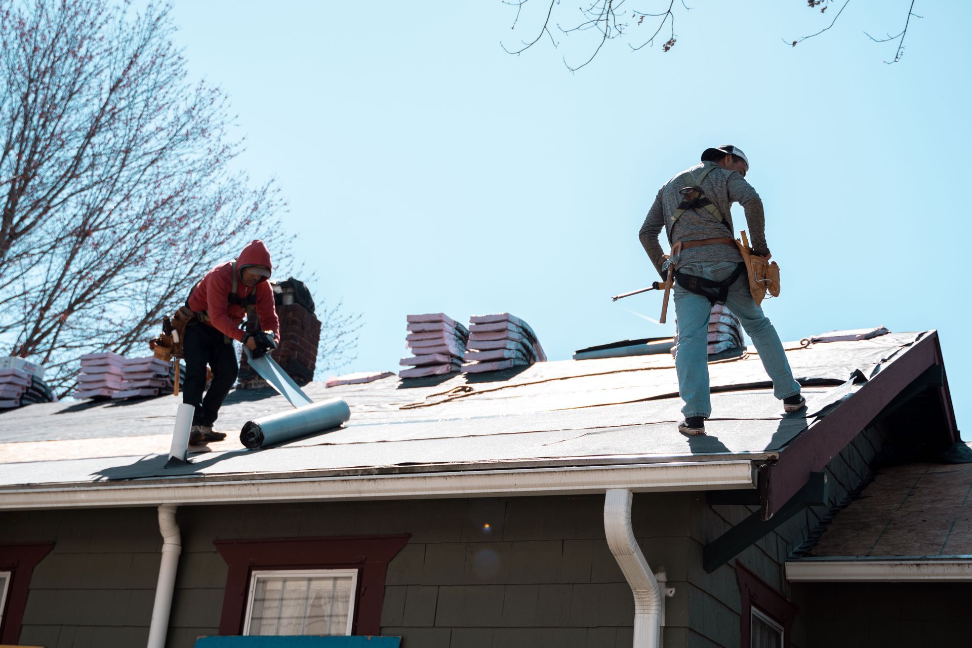 Two roofers working on a roof, bundles of shingles nearby, clear sky overhead.
