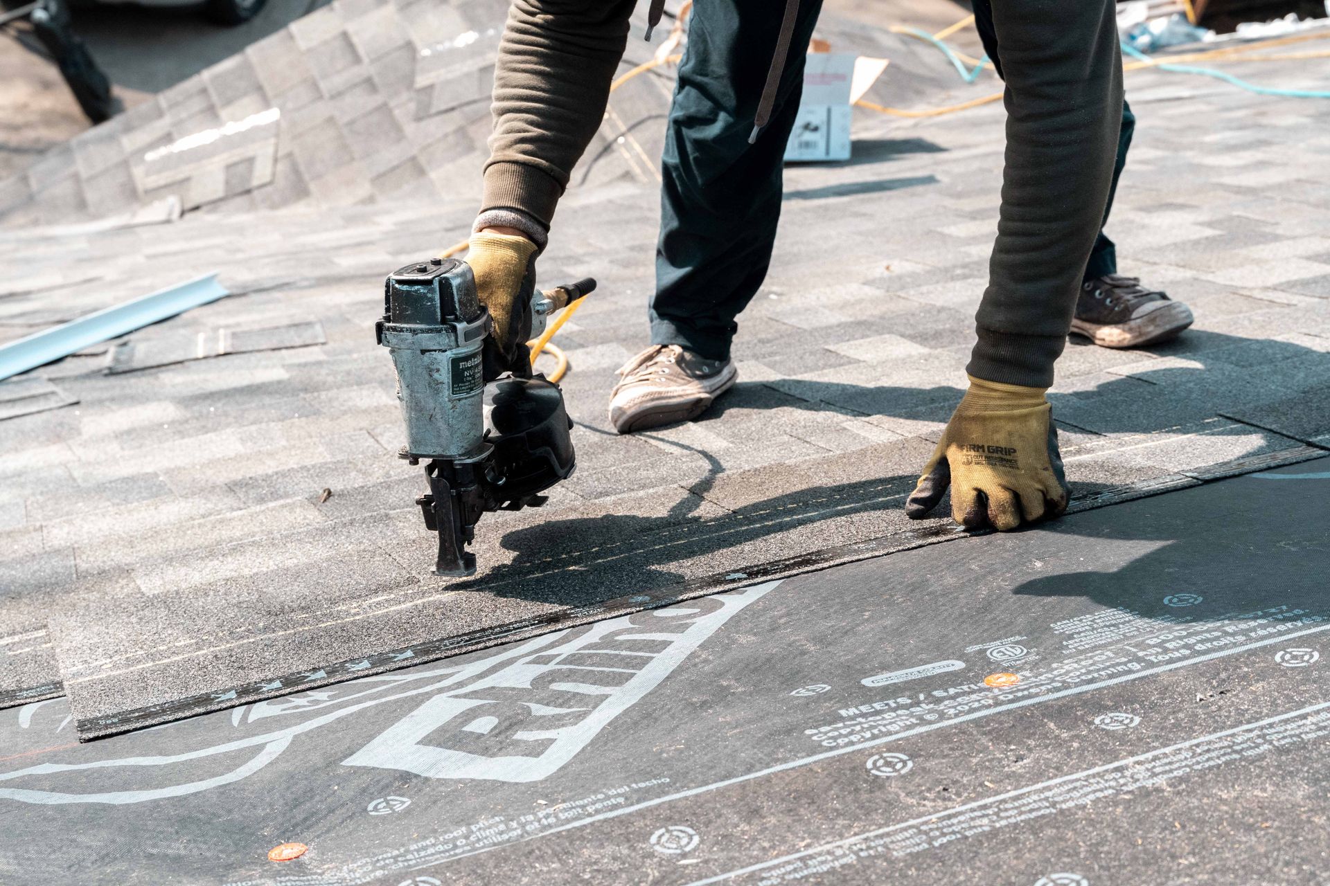 Roofer using a nail gun to secure roofing material on a dark shingled roof, outdoors.