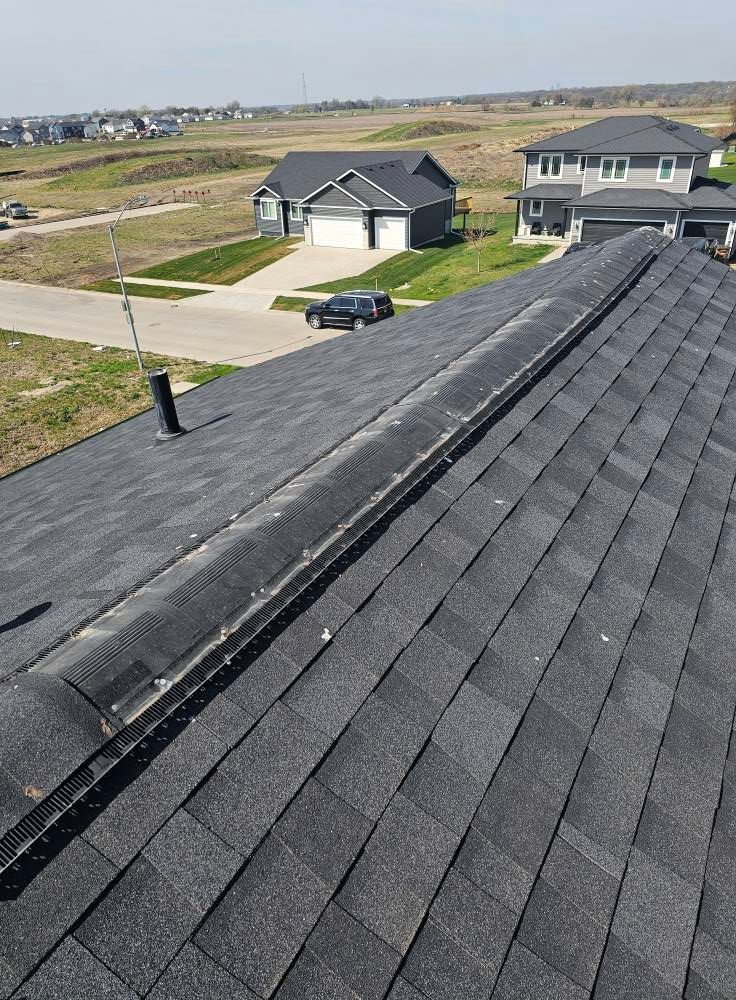 Black shingle roof with a vent, overlooking suburban houses and landscape on a sunny day.