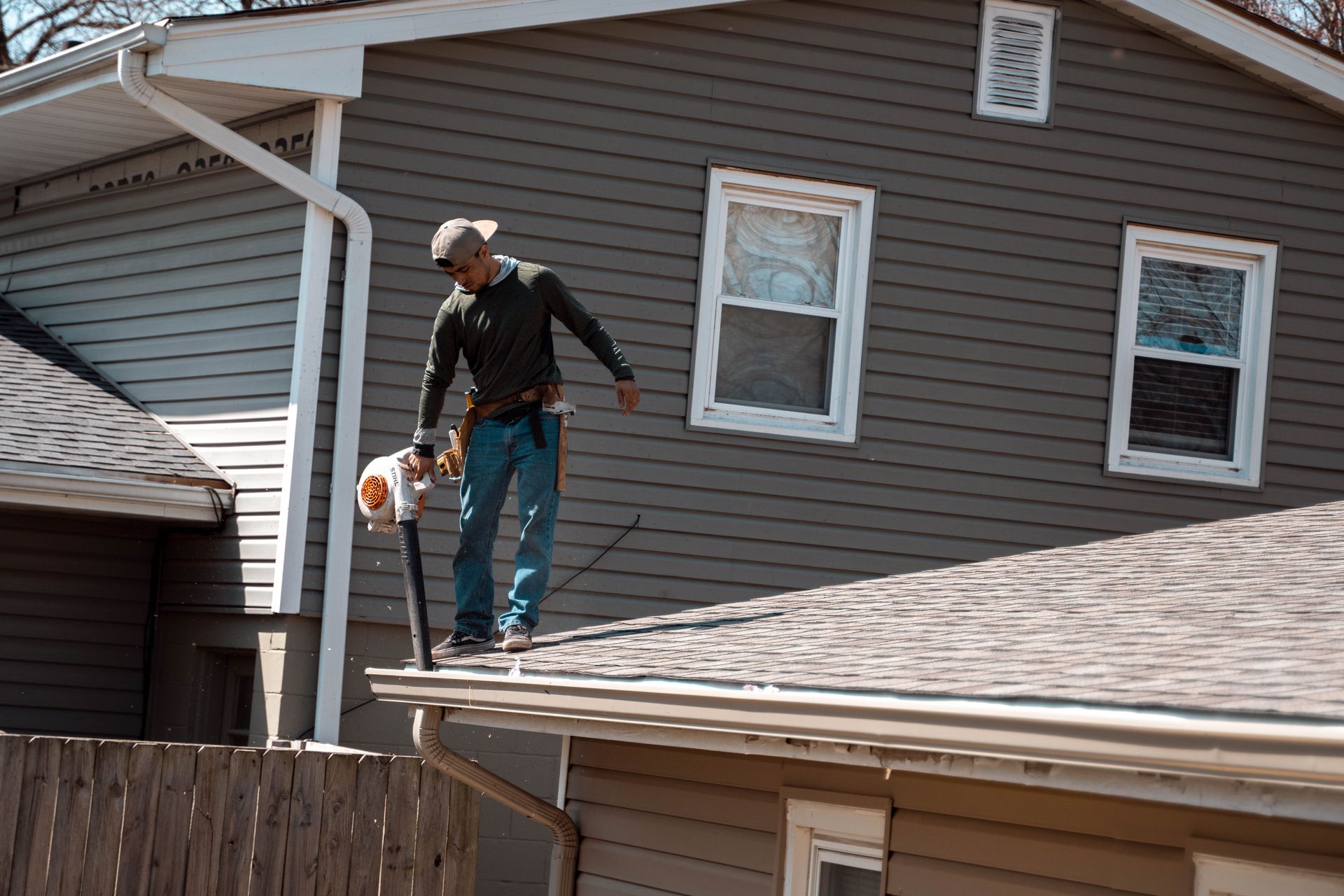 Man on a roof using a leaf blower to clean gutters on a gray house.