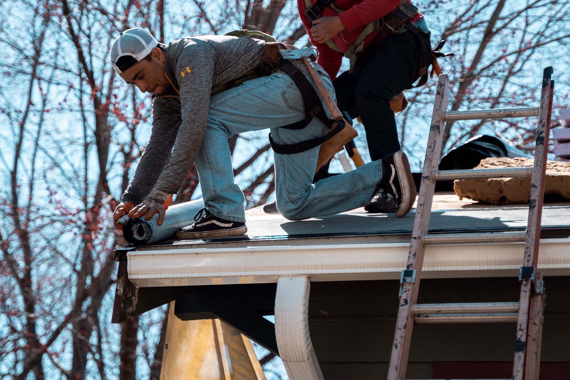 Two workers installing roofing material on a house; one rolls out material while another stands behind.