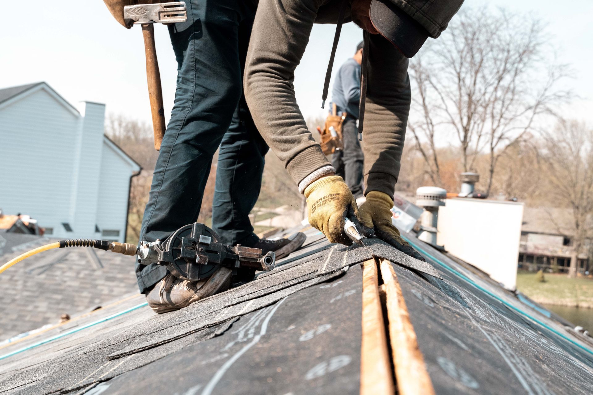 Roofer installing shingles on a house roof, holding materials and hammer in sunny conditions.