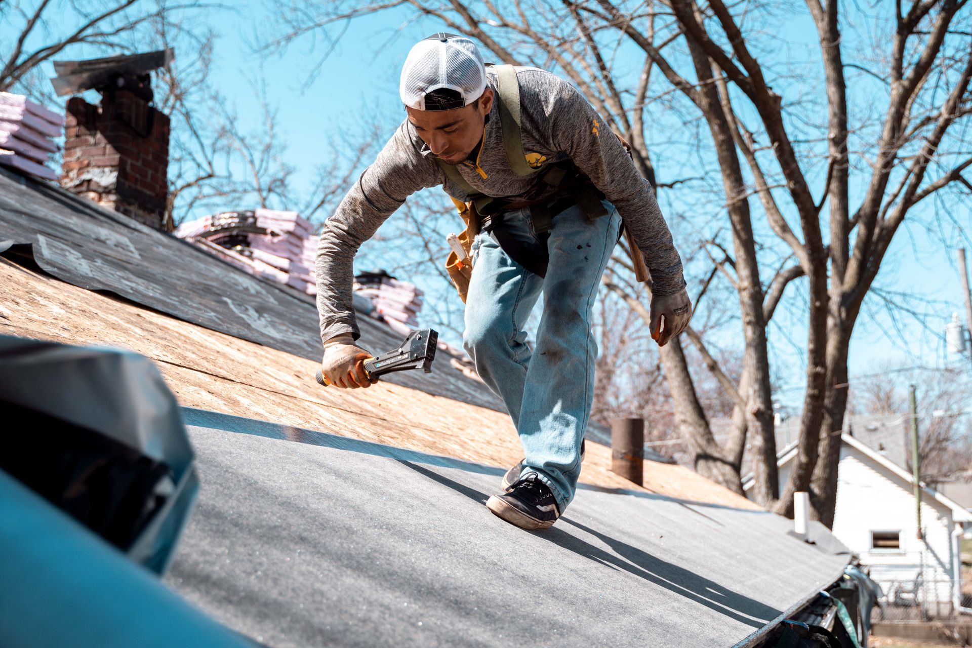 Roofer works on a house, hammer in hand. Blue jeans, camo shirt, white hat. Sunny day.