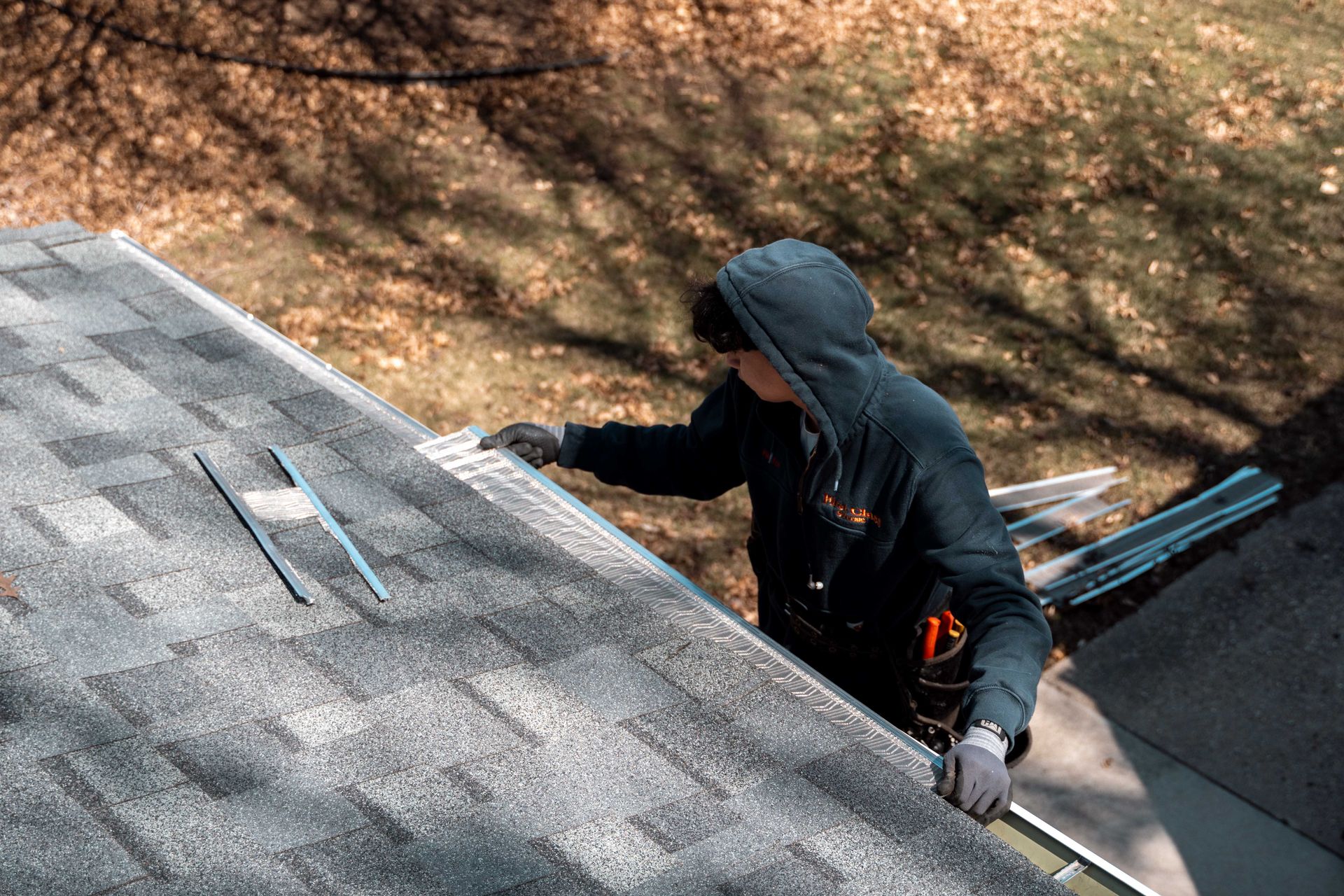 Person on a rooftop, wearing a hooded sweatshirt and gloves, working on a shingled roof with a gutter nearby.
