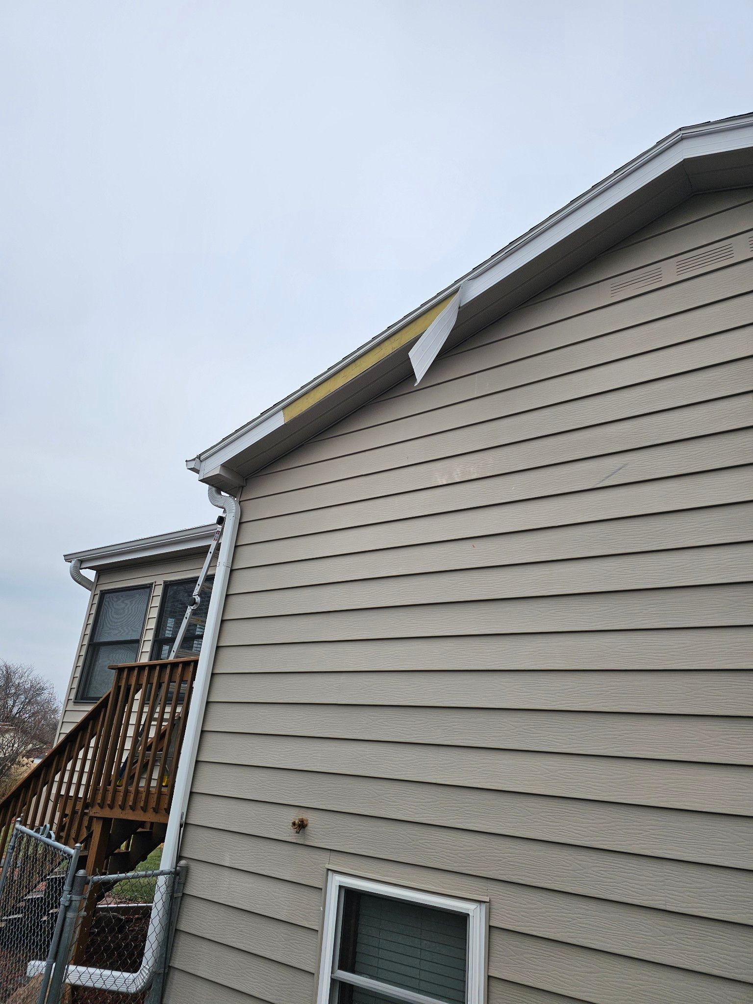 Side view of a house with light tan siding and a detached piece of yellow material near the roof.