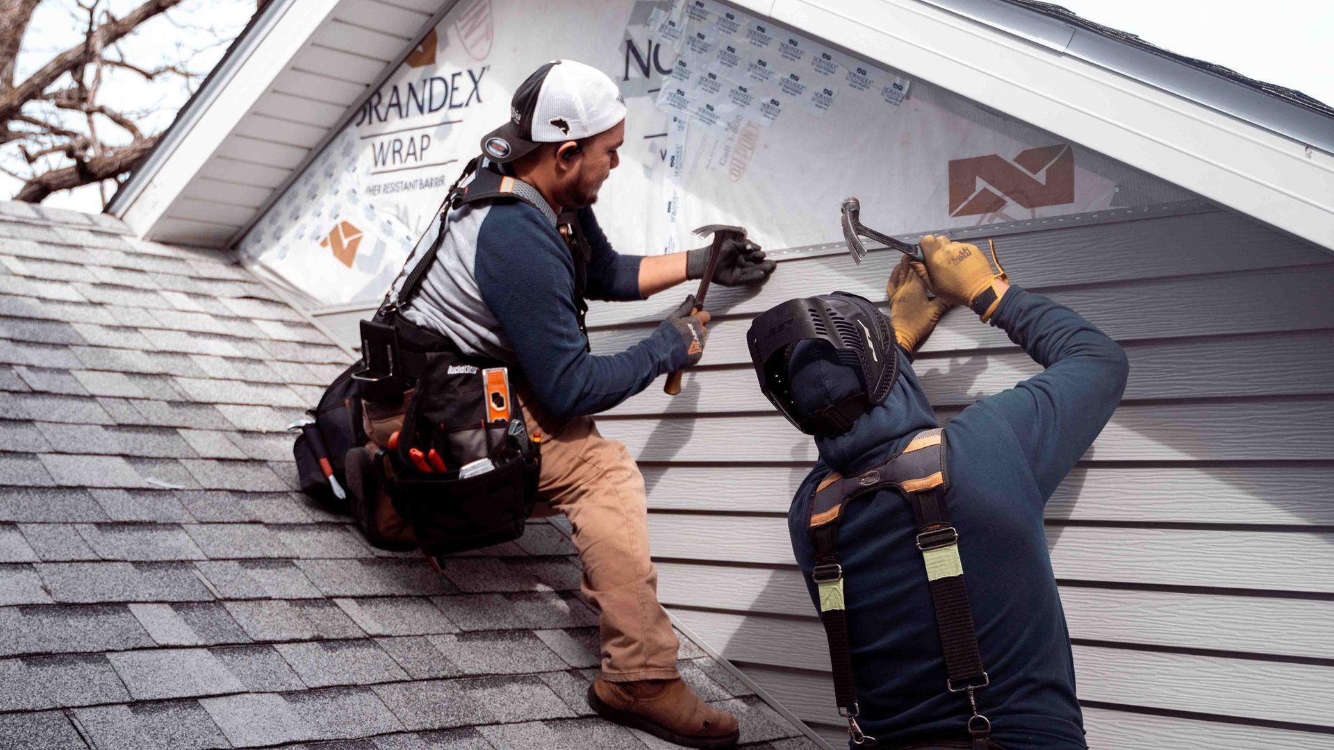 Two workers install siding on a roof. One uses a hammer, the other holds a panel, both wear safety gear.