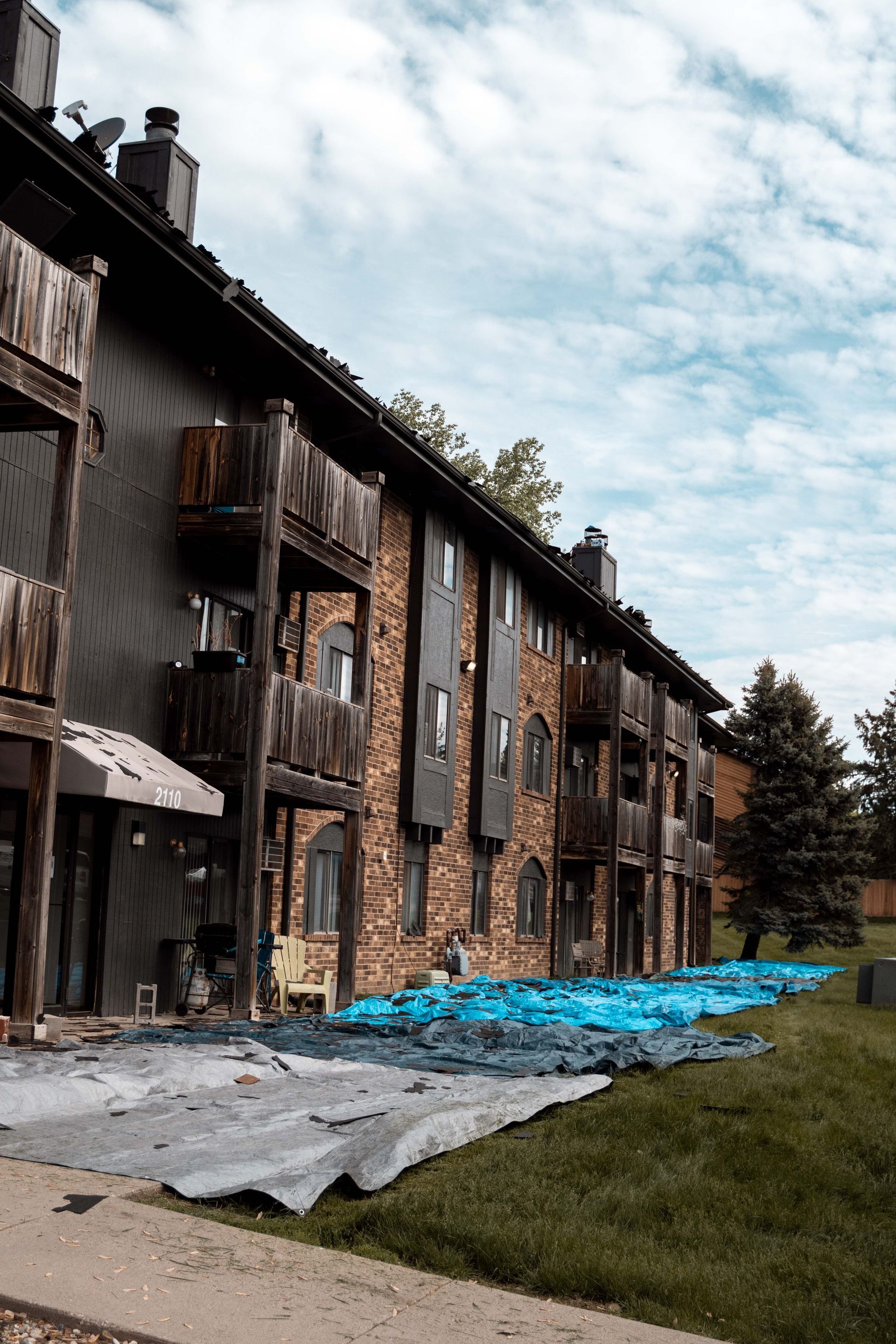 Apartment building with blue tarp covering the ground. Brown brick and siding, balconies, cloudy sky.