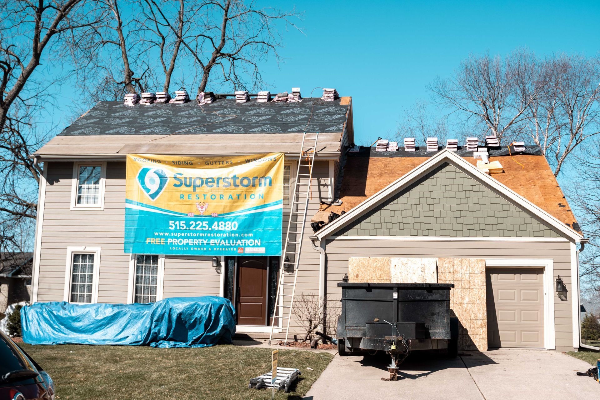 House with roofing partially removed, blue tarp, Superstorm Restoration banner, and trash bin.
