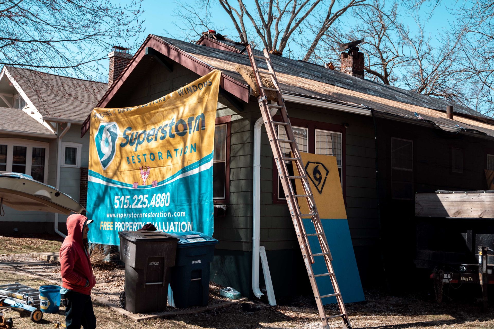 A house with roofing work in progress; a person in a red hoodie stands near a dumpster; a ladder leans against the building.