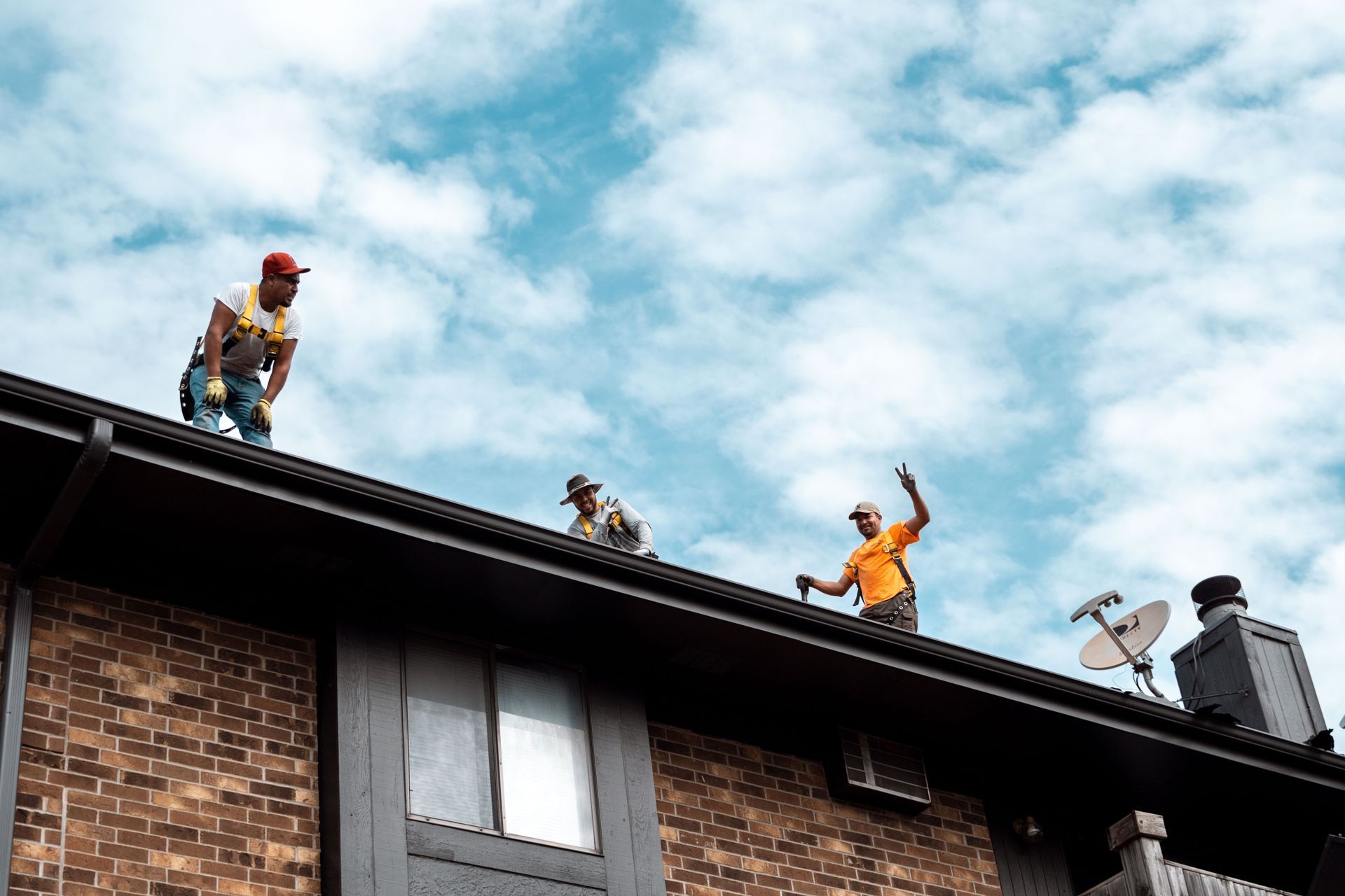 Three workers on a roof repair gutters; cloudy sky above a brick building.