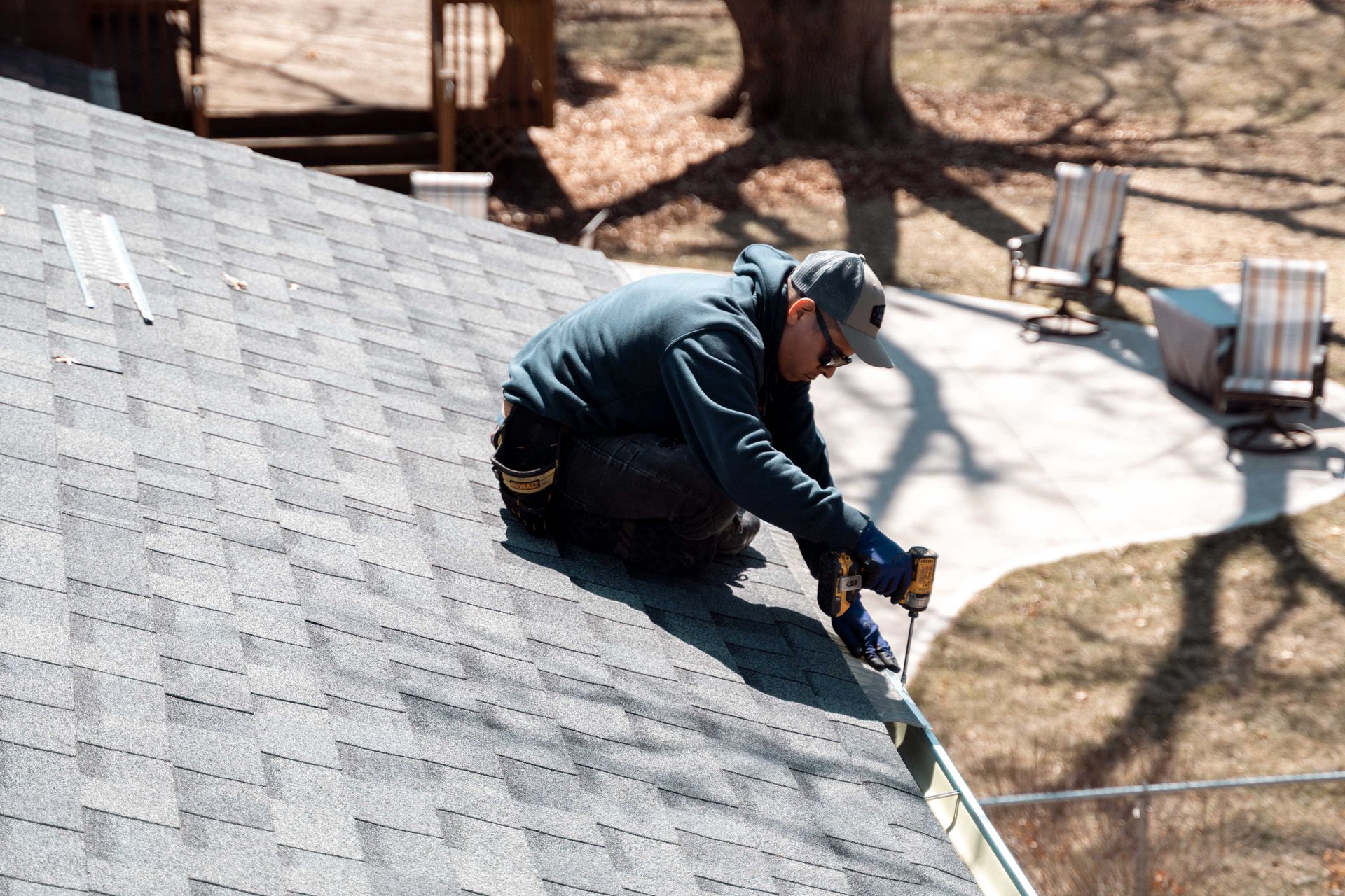 Roofer installing a gutter on a residential home. Blue shirt, gray roof, sunny day.