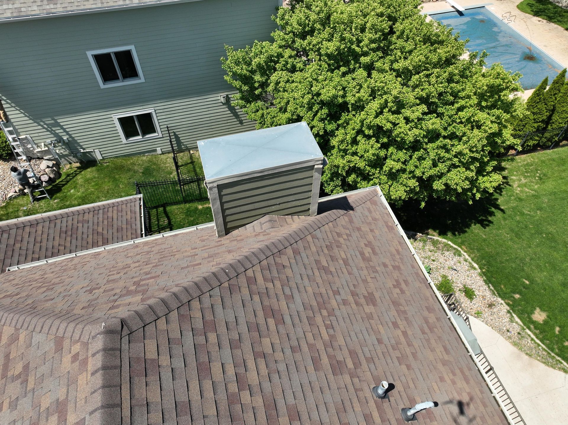 Overhead view of a house roof, chimney, and adjacent green lawn with a swimming pool and tree.