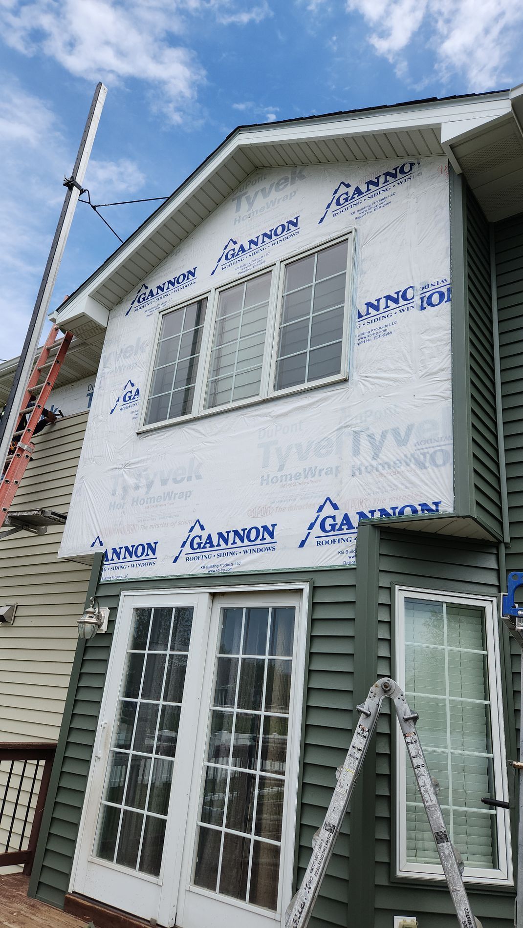 Two-story house with green siding, Tyvek wrap, windows. A ladder and scaffolding are present.